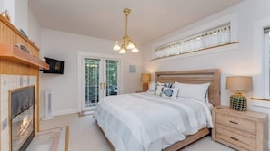 A bedroom with a large bed, a wooden headboard, a TV mounted on the wall, a fireplace, and a window with curtains.