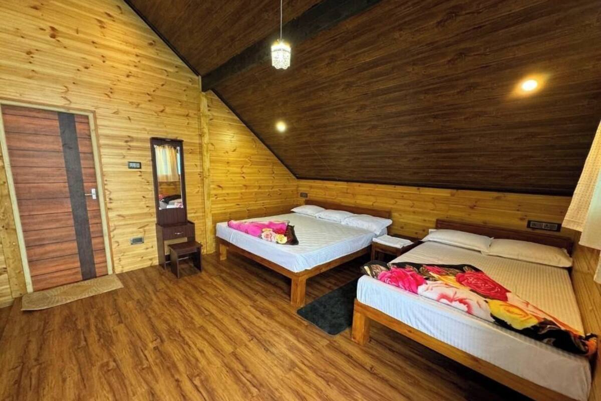 Une pièce aux murs en bois et au plafond en pente. On y trouve deux lits avec du linge de lit blanc et des couvertures colorées. Une petite table en bois et une chaise sont placées près de l  ’un des lits. Une porte avec un miroir et un interrupteur sont visibles.
