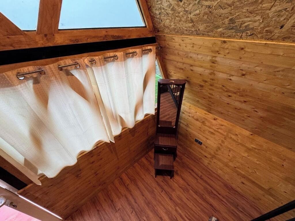Un angolo di stanza con pareti in legno, una finestra con tenda e un piccolo sgabello di legno.