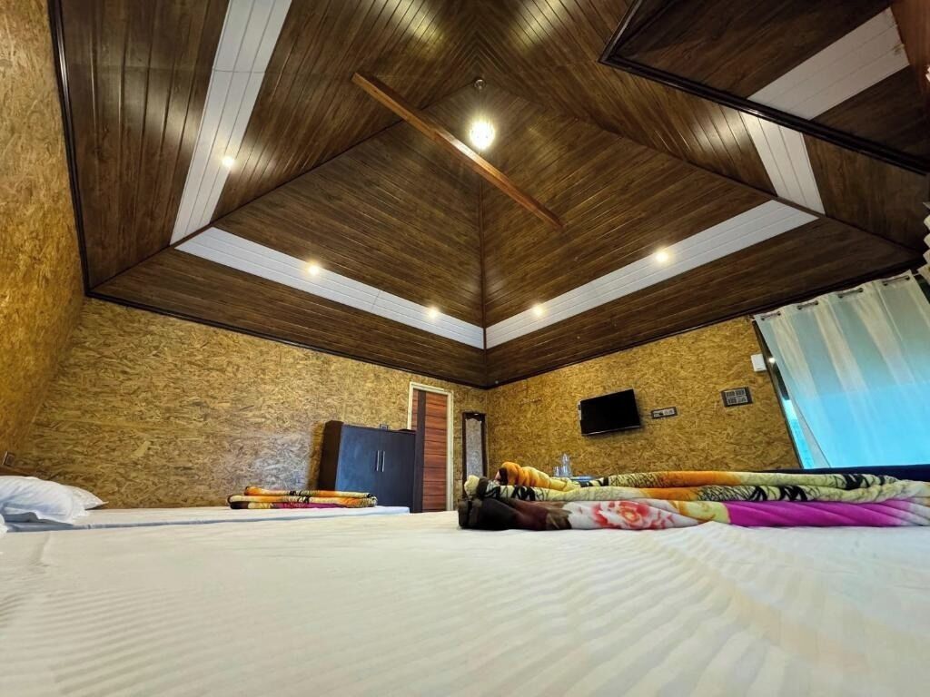 Ein Zimmer mit hoher Decke, Holzwänden, einem Bett mit weißer Matratze, einem Kleiderschrank, einem Flachbildfernseher und einem Fenster mit Vorhängen.