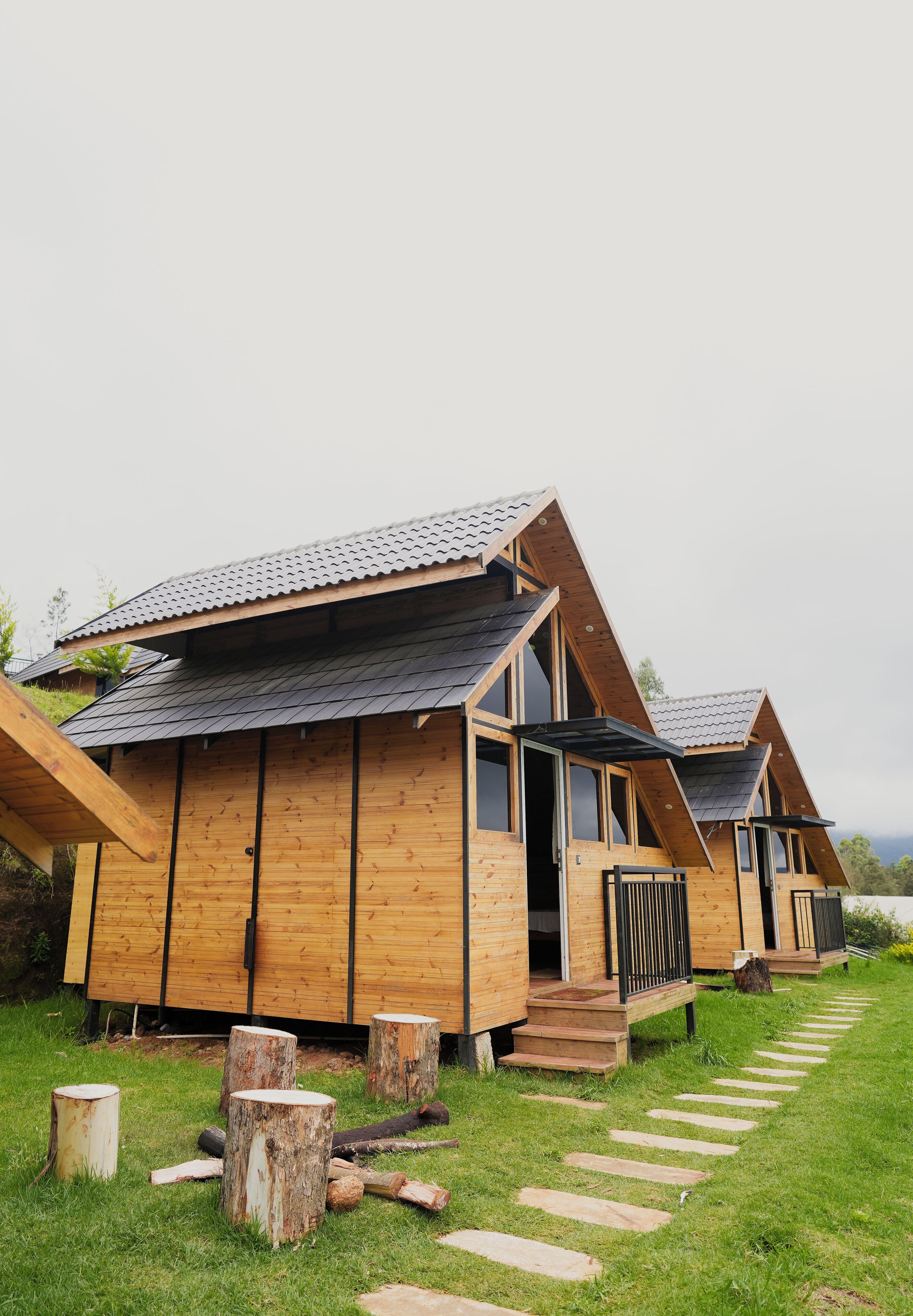 A row of wooden cabins with pitched roofs and large windows, set on a grassy path with wooden stumps.