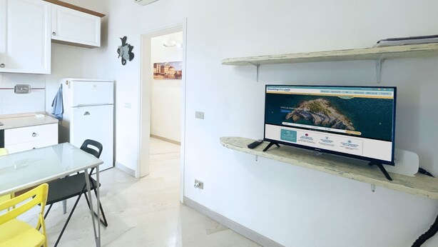 A modern kitchen with white cabinets, a refrigerator, a dining table with chairs, and a mounted flat-screen TV displaying a scenic view. - Casa Elia - Procchio - Casa Elia - Procchio (Marciana)