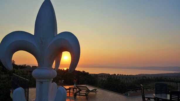 Une vue sur le coucher de soleil depuis un balcon agrémenté d’une grande sculpture en forme de fleur de lys, avec un espace salon extérieur et une vue sur le paysage lointain.