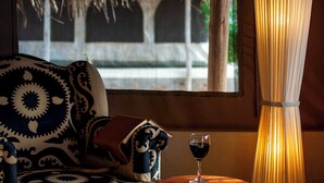 A room with a patterned sofa, a small round table with a glass of red wine, and a tall, pleated lamp. - Kudu Safari Camp (Tsavo East)