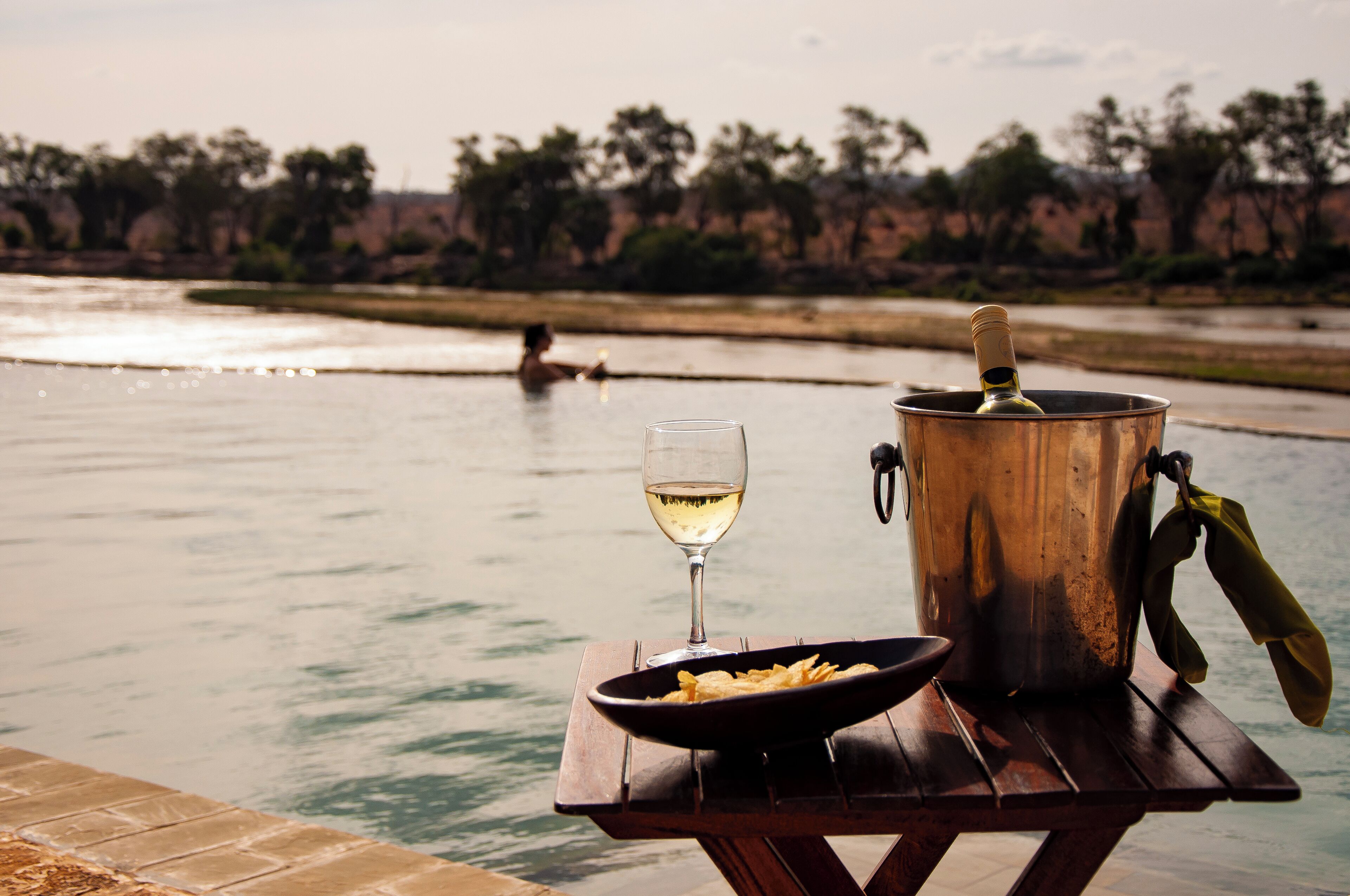 Een houten tafel aan de waterkant met een champagnefles, een glas en een schaal chips.