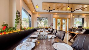 A restaurant with a long dining table set for a meal, surrounded by leather chairs, with a view of the outdoors through large windows. -  Wonderwood Corbett Resort (1min walk to River with Deep Jungle Trail) (Ramnagar)