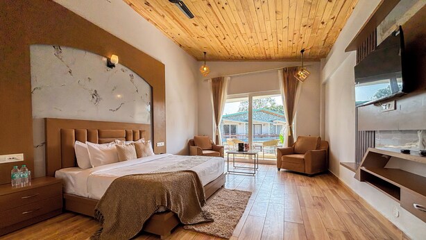A modern hotel room with a wooden ceiling, a large bed, a seating area with two chairs, a TV, and a view of the outside. -  Wonderwood Corbett Resort (1min walk to River with Deep Jungle Trail) (Ramnagar)