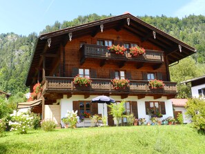 A traditional wooden chalet with multiple balconies, surrounded by lush greenery and a mountainous backdrop.