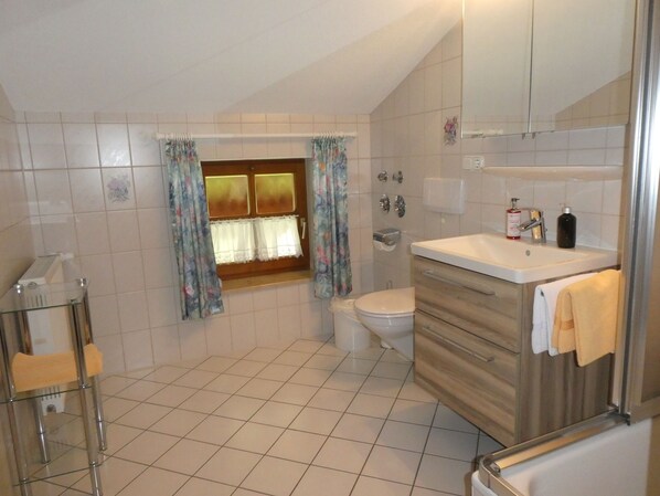 A bathroom with a toilet, sink, and shower. There is a window with curtains, a towel hanging on the wall, and a glass table with a chair.
