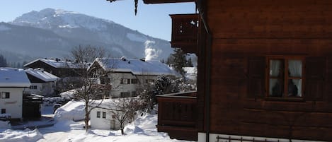 Un edificio de madera con techo nevado, una ventana con cortina y vista a una montaña al fondo.
