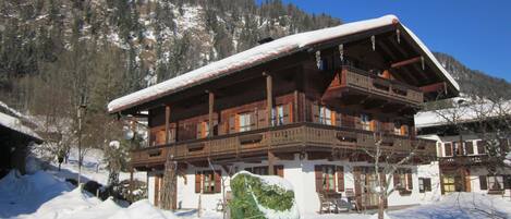 A wooden chalet with a balcony, surrounded by snow-covered ground and trees.