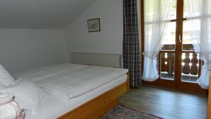 A bedroom with a wooden bed, a radiator, a patterned rug, and a view through a door with curtains. - Apartment (03) with balcony and extra kitchen (Reit im Winkl)