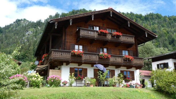 A traditional wooden chalet with balconies adorned with flowers, surrounded by lush greenery and a mountainous backdrop. - Apartment (03) with balcony and extra kitchen (Reit im Winkl)