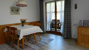 A dining area with a table set for two, wooden chairs, a vase with flowers, a patterned rug, and a view through a window with curtains. - Apartment (03) with balcony and extra kitchen (Reit im Winkl)