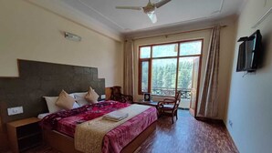 A bedroom with a large bed, a desk, and a chair. There is a window with curtains, a ceiling fan, and a television on the wall. - Chichoga Holiday Inn Manali (Manali)