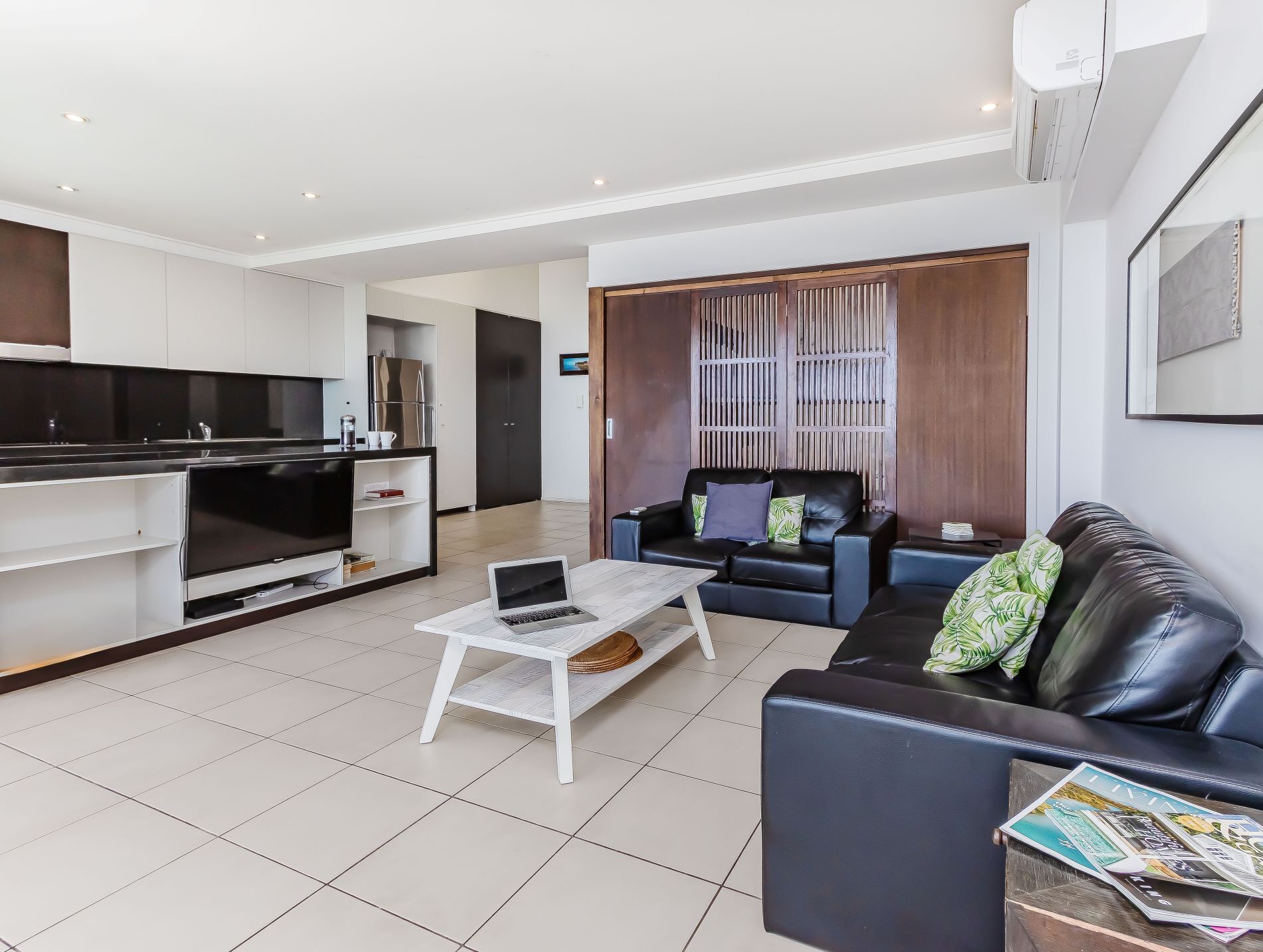 Una sala de estar moderna con un sofá de cuero negro, una mesa de centro blanca y un sofá cama de cuero negro. Hay una zona de cocina con armarios blancos y una encimera negra.