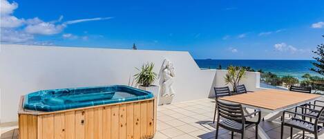 Una bañera de madera en un patio con baldosas, con una mesa y sillas, con vistas a una playa y al océano.