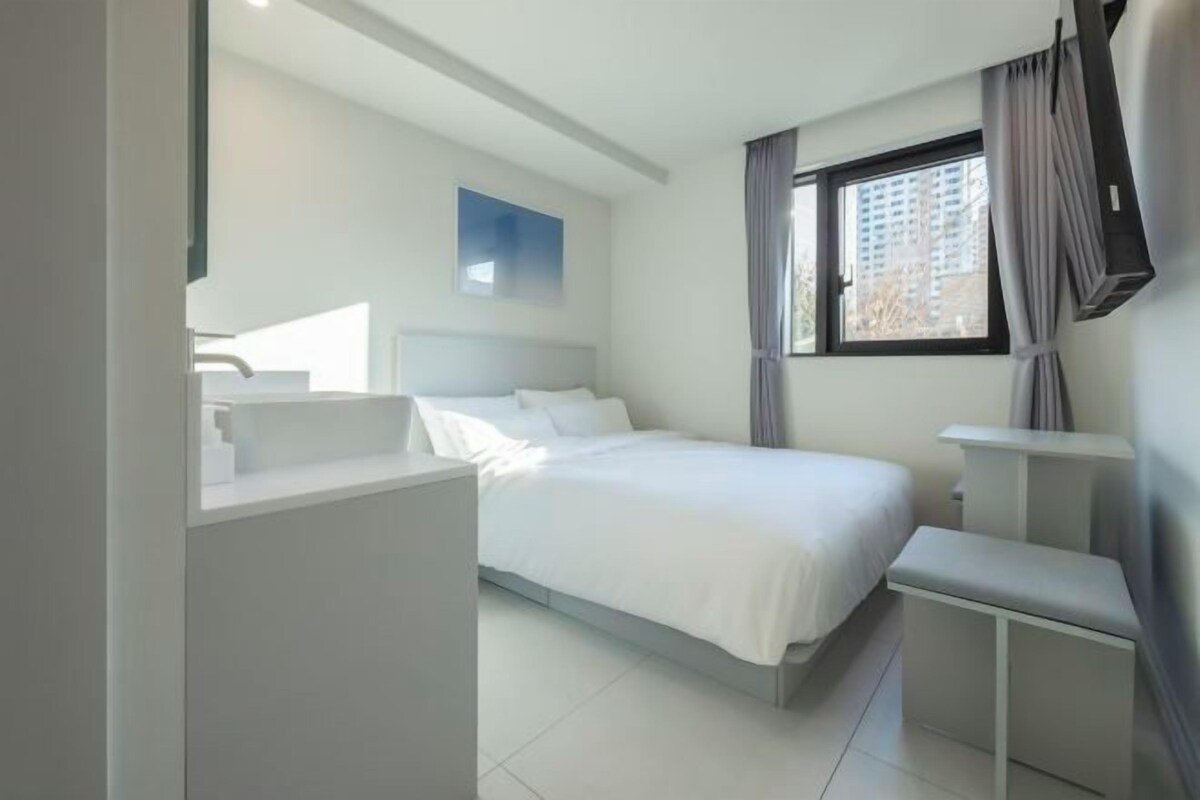 A modern hotel room with a large bed, a bedside table, a small bench, and a window with curtains.