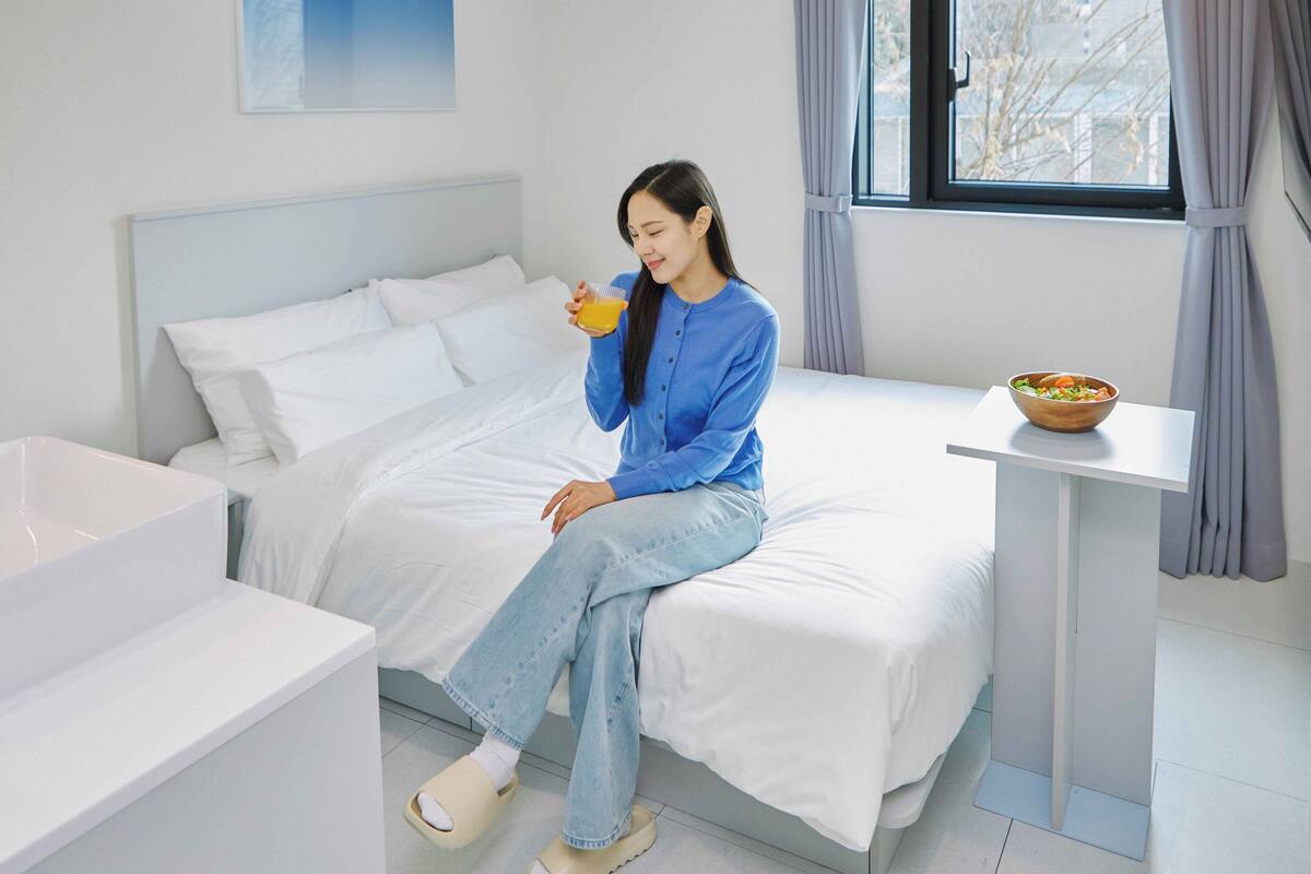 Une personne est assise sur un lit dans une chambre d’hôtel. On aperçoit un couvre-lit blanc, une petite table avec un saladier rempli de fruits et une fenêtre avec des rideaux.