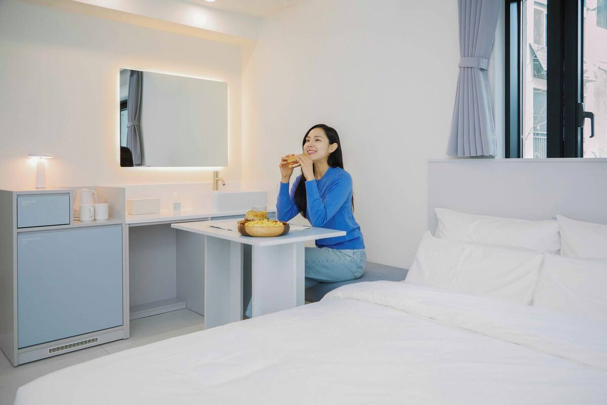 Une chambre d’hôtel avec un lit, une table de chevet sur laquelle se trouve un bol de nourriture, un petit bureau avec une lampe, un miroir et une fenêtre avec des rideaux.
