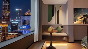 Habitación de hotel moderna con vistas a la ciudad, una mesa redonda, un sofá con cojines y un cuadro grande en la pared.