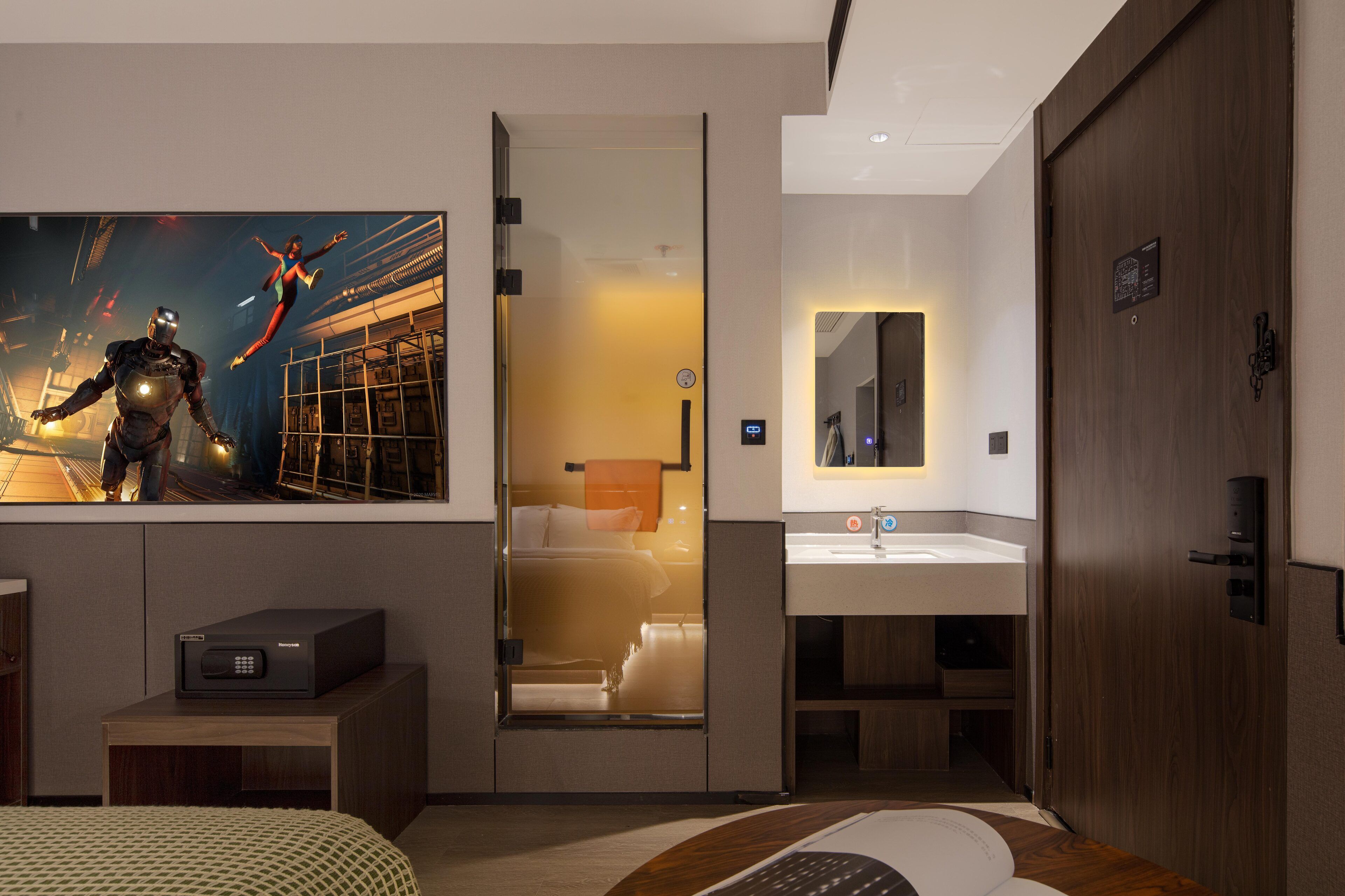 Habitación de hotel moderna con una cama grande, un escritorio, un televisor mostrando una escena de superhéroes, un baño con espejo y lavamanos, y un armario de madera.