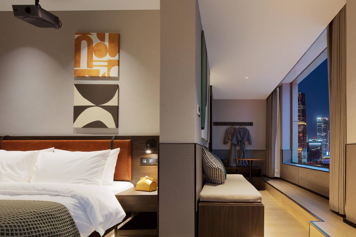 Quarto de hotel moderno com uma cama grande, uma escrivaninha e vista para a cidade à noite.