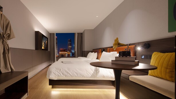 Una habitación de hotel moderna con una cama grande, una mesita de noche, una pequeña mesa redonda y vistas a un paisaje urbano nocturno.
