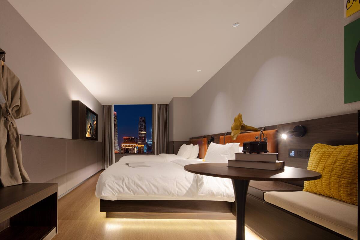 Quarto de hotel moderno com uma cama grande, mesa de cabeceira, pequena mesa redonda e vista para o horizonte noturno.