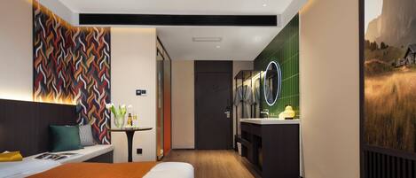 A modern hotel room with a bed, a small table with a vase, a mirror, and a bathroom with a sink and toilet.