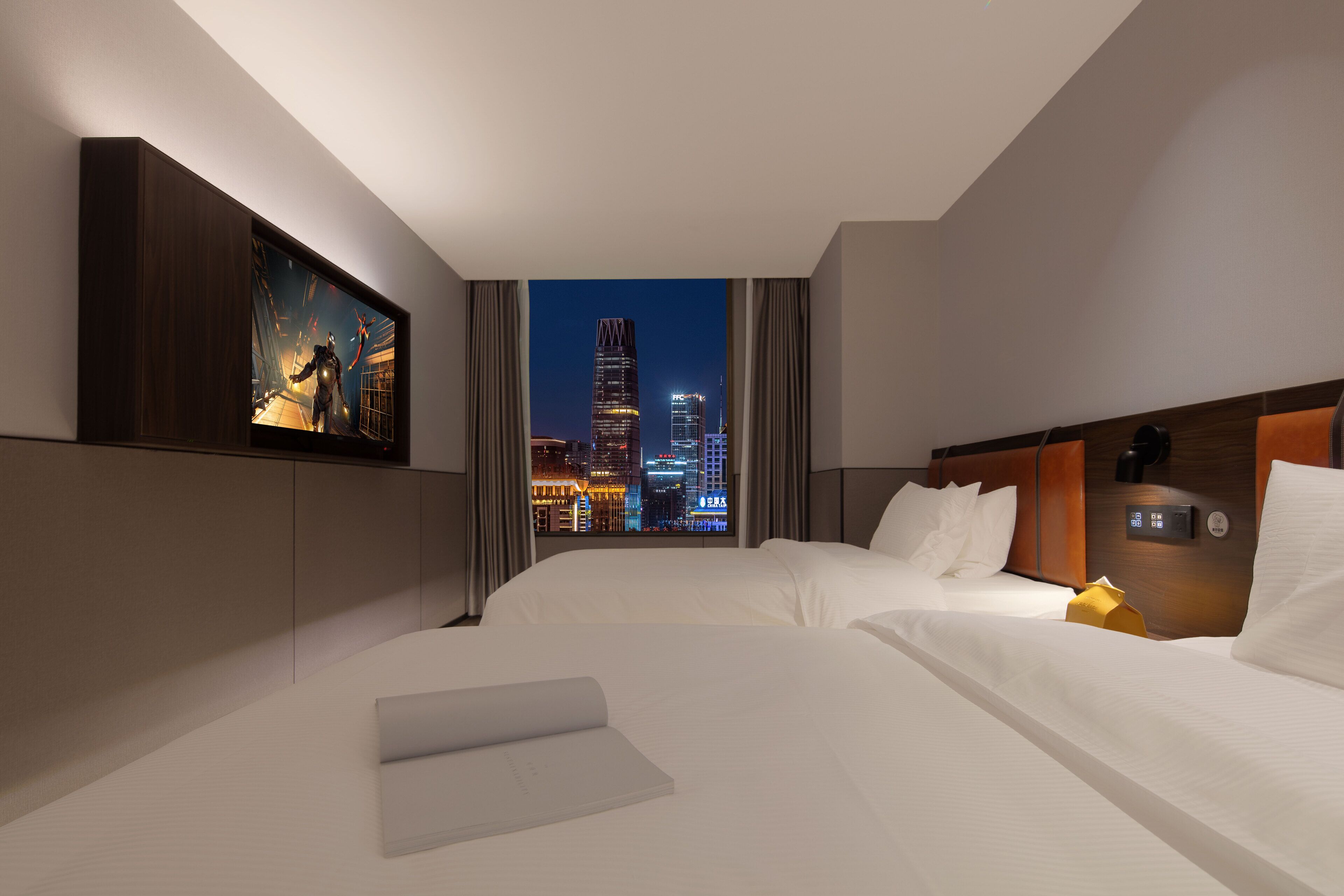A hotel room with two beds, a city view, a painting on the wall, and a modern control panel.