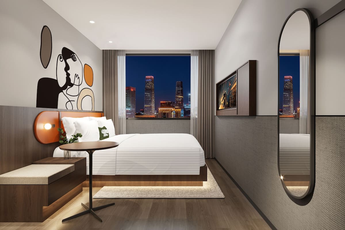 Quarto de hotel moderno com cama, mesa de cabeceira e uma janela grande com vista para a cidade.