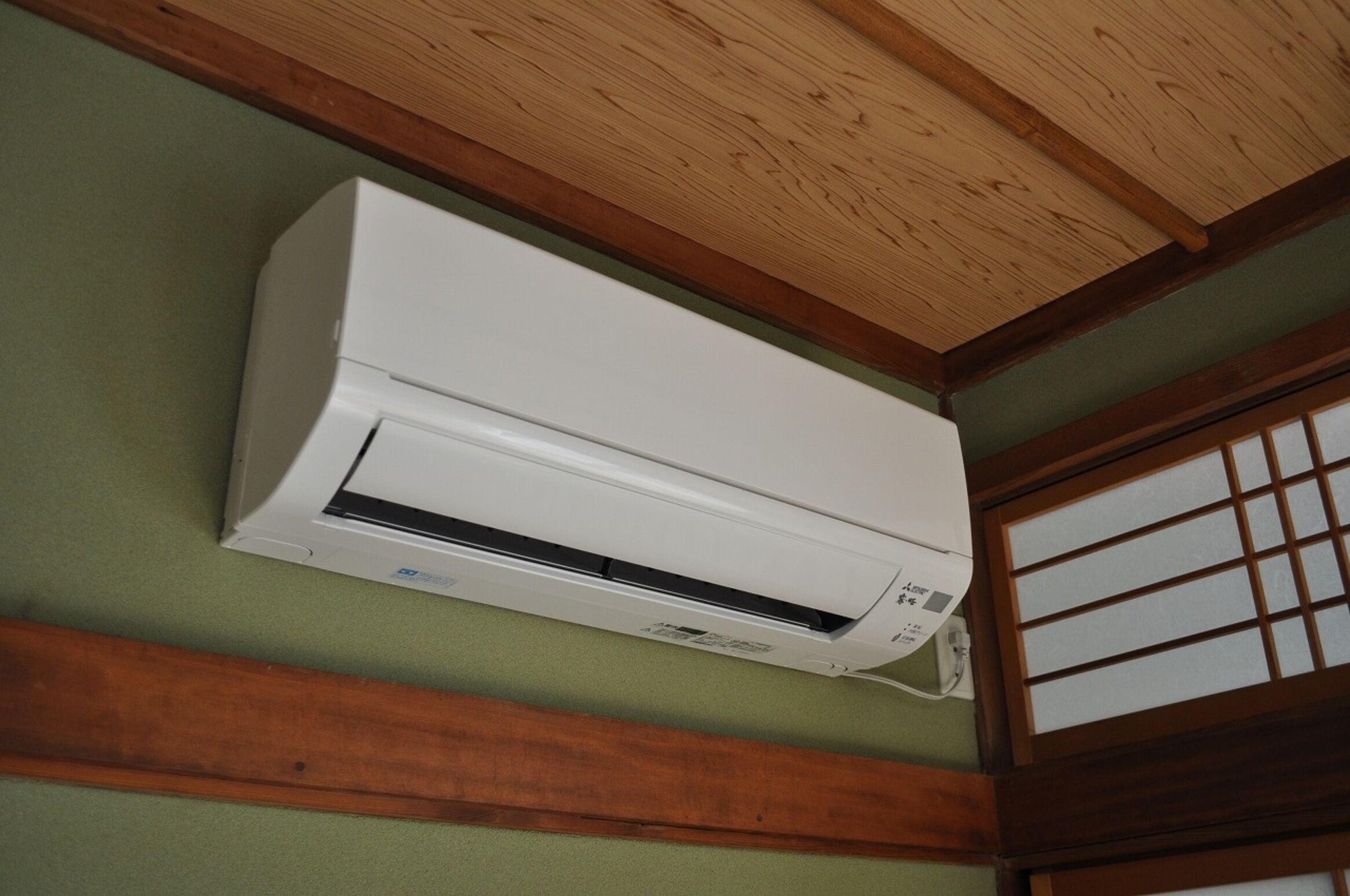 A white air conditioning unit mounted on a green wall with wooden beams and a traditional window with grid panes.