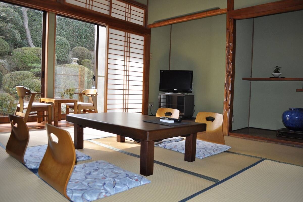 A traditional Japanese room with tatami mats, a low table, wooden chairs, a television, and a view of a garden through shoji screens.