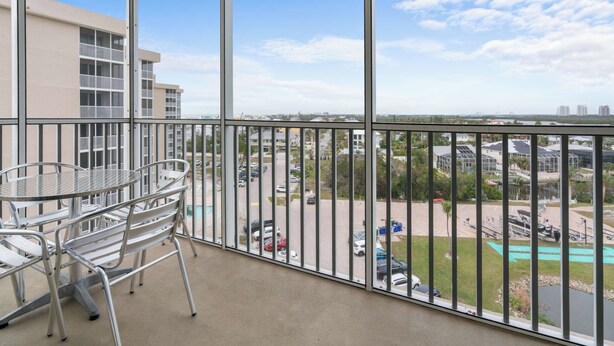 A balcony with a glass table and chairs, overlooking a parking area and buildings.
