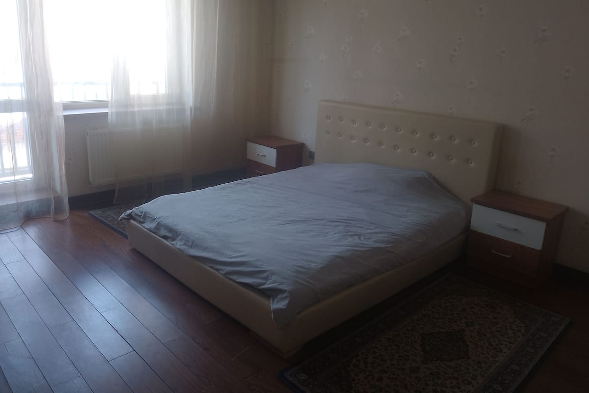 A bedroom with a bed, two nightstands, a window with sheer curtains, and a radiator.