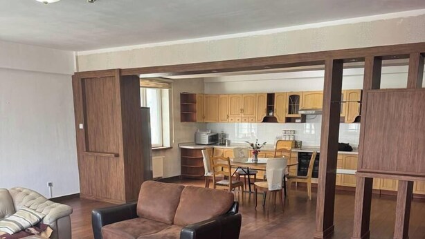A spacious living room with a brown leather sofa, wooden coffee table, and a dining area with a white table and chairs. A kitchen with wooden cabinets and a window is visible in the background. - Guest House Ulaanbaatar (Ulaanbaatar)