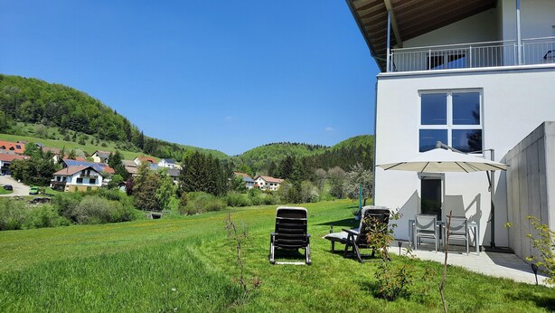 Een modern huis met een balkon, een gazon met stoelen en uitzicht op de omliggende heuvels.
