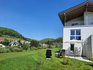 A modern house with a balcony, a lawn with chairs, and a view of the surrounding hills.