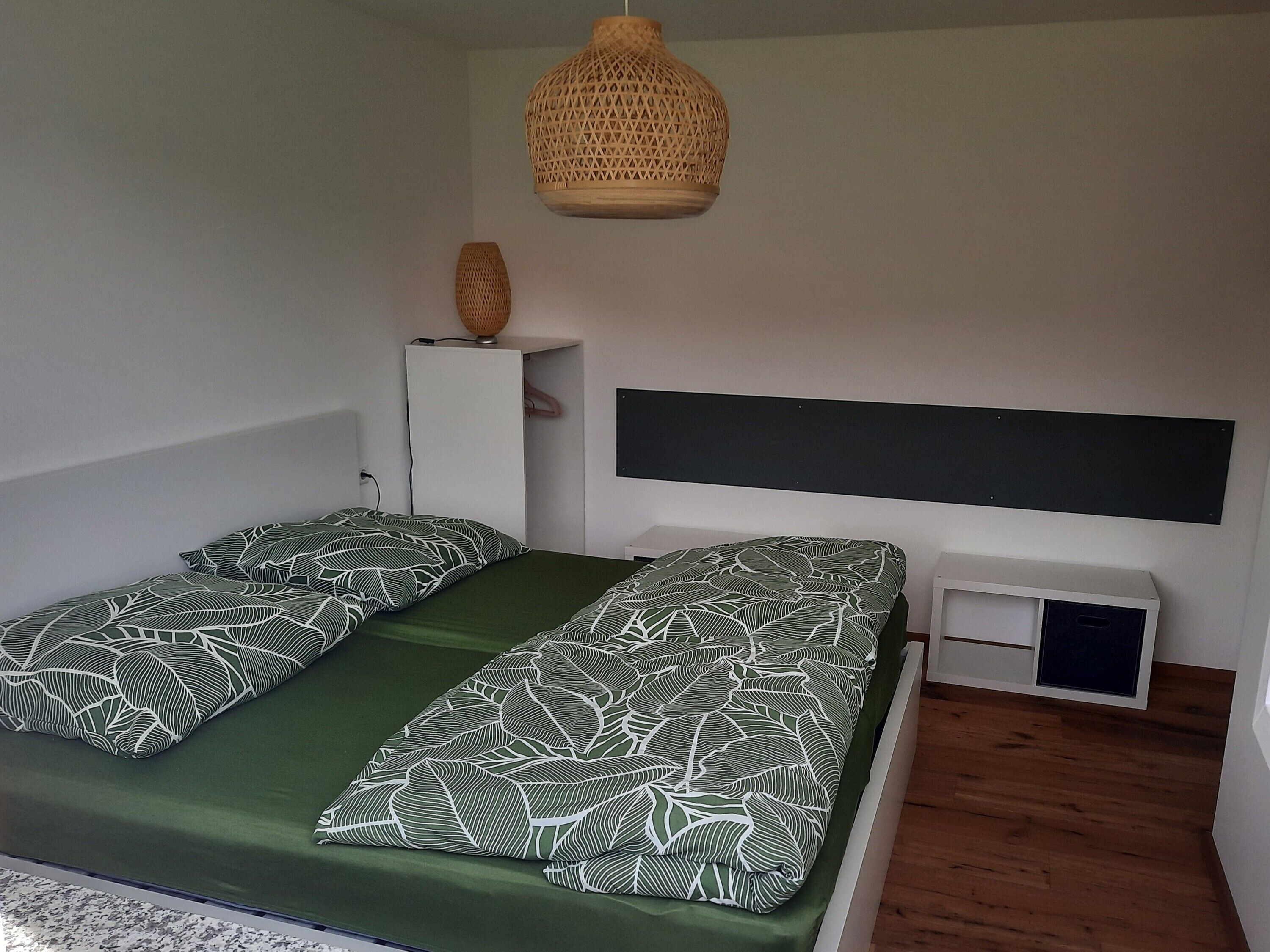 Een kamer met een bed, een wit kastje, een geweven lamp en een zwart hoofdeinde.