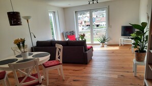Een woonkamer met een bank, eettafel, stoelen, een televisie en een groot raam met uitzicht op gebouwen.