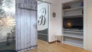 Ein modernes Wohnzimmer mit großem Fenster, einer Holztür mit 'B'-Beschriftung, einer weißen TV-Konsole und einem kleinen weißen Tisch.