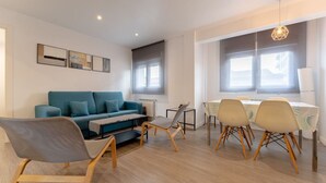 Ein modernes Wohnzimmer mit einem blauen Sofa, Holzstühlen, einem Esstisch mit weißer Tischdecke und einem großen Fenster mit Jalousien.