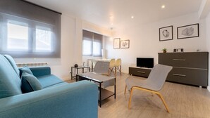Ein modernes Wohnzimmer mit einer Couch, einem Esstisch und einem Fernseher. Zwei Fenster mit Jalousien, ein Schrank und eine Pflanze sind vorhanden.