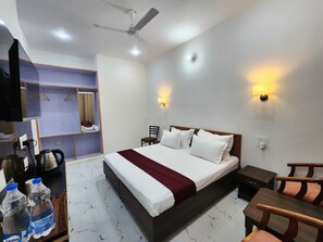 A hotel room with a bed, a desk, a chair, a wardrobe, a ceiling fan, and wall-mounted lights. - Dorjee House & Rooftop Cafe (Baijnath)