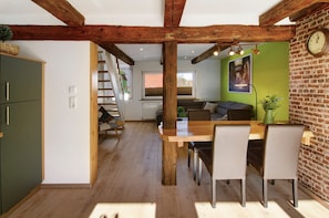 Un coin repas avec une table et des chaises en bois, un mur vert orné d’une horloge, et un escalier menant à une mezzanine.