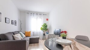 Una sala de estar moderna con un sofá gris, una mesa de comedor con platos y vasos, una planta en maceta y un televisor.