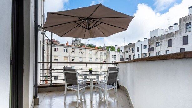 Ein Balkon mit Tisch und Stühlen, einem großen Sonnenschirm und Blick auf umliegende Gebäude.