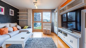 Ein modernes Wohnzimmer mit Sofa, Couchtisch und Fernseher. Ein großes Fenster bietet Blick auf Gebäude und Bäume.