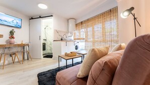 Ein modernes Wohnzimmer mit Sofa, einem Esstisch mit Stühlen, einer Küchenzeile inklusive Waschmaschine und einem großen Fenster mit Jalousien.